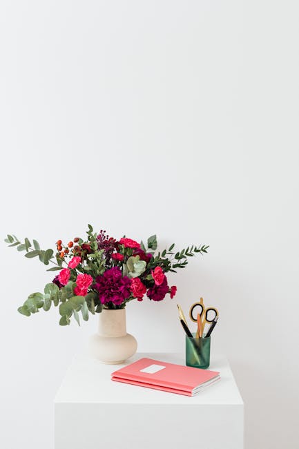 A floral arrangement featuring vibrant pink and deep red carnations, complemented by sprigs of grey-green eucalyptus leaves, is displayed in a simple, off-white ceramic vase on a white surface. The bo