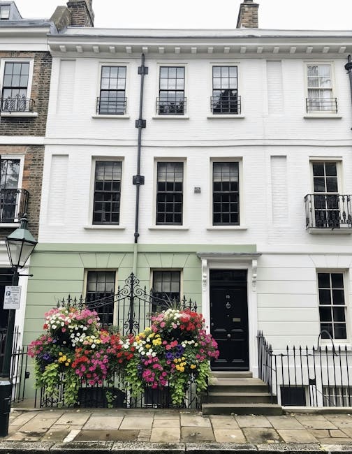 A vibrant floral display featuring two large, lush bouquets of mixed seasonal flowers positioned on either side of a black wrought iron gate in front of a white, multi-storey residential building. The