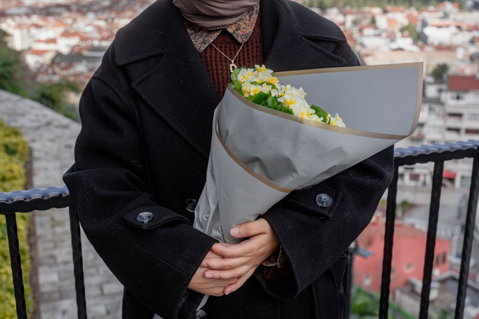 A person dressed in a black coat holding a bouquet of fresh flowers wrapped in light grey paper with a beige border, featuring predominantly white and yellow blossoms, possibly daisies or chrysanthemu