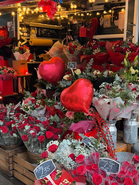 A floral display at a flower shop featuring an arrangement of fresh red and white roses, carnations, and greenery, with two large red heart-shaped balloons among the bouquets. The flowers are wrapped 