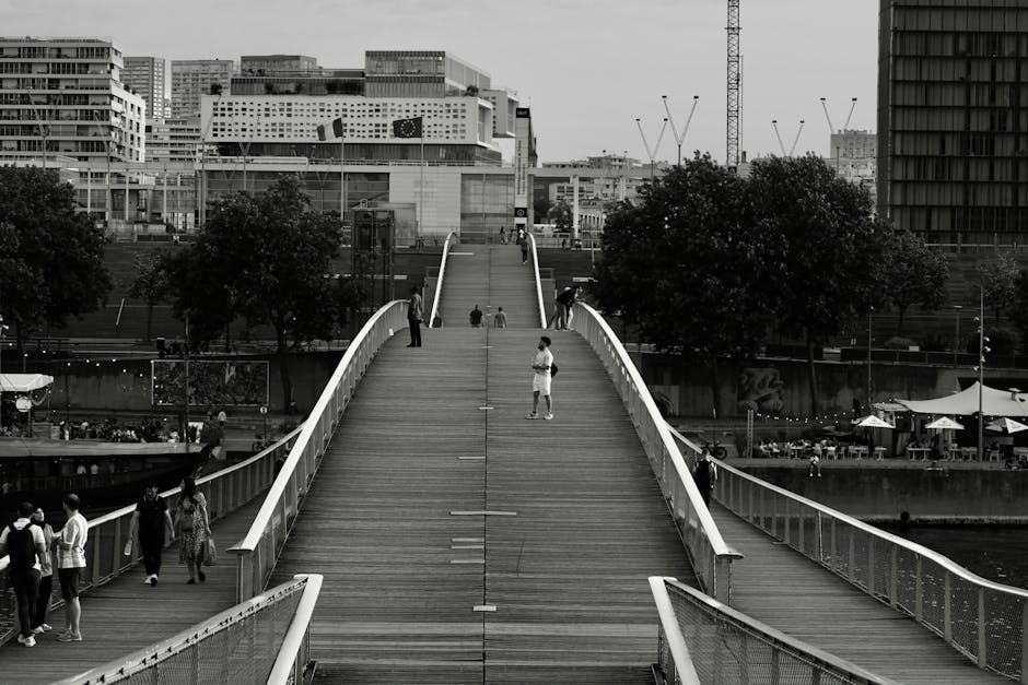 A black-and-white photograph of a modern pedestrian bridge with a gently curved wooden surface and white metal railings, spanning over a waterway in an urban setting. Several people are walking or sta