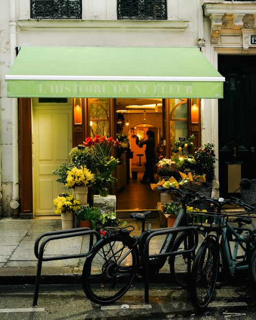 View of the florist shop 'L'Histoire du Fleurs' with a bright green awning displaying the shop's name, showcasing a display of fresh flower bouquets in various vases and buckets arranged outside the e
