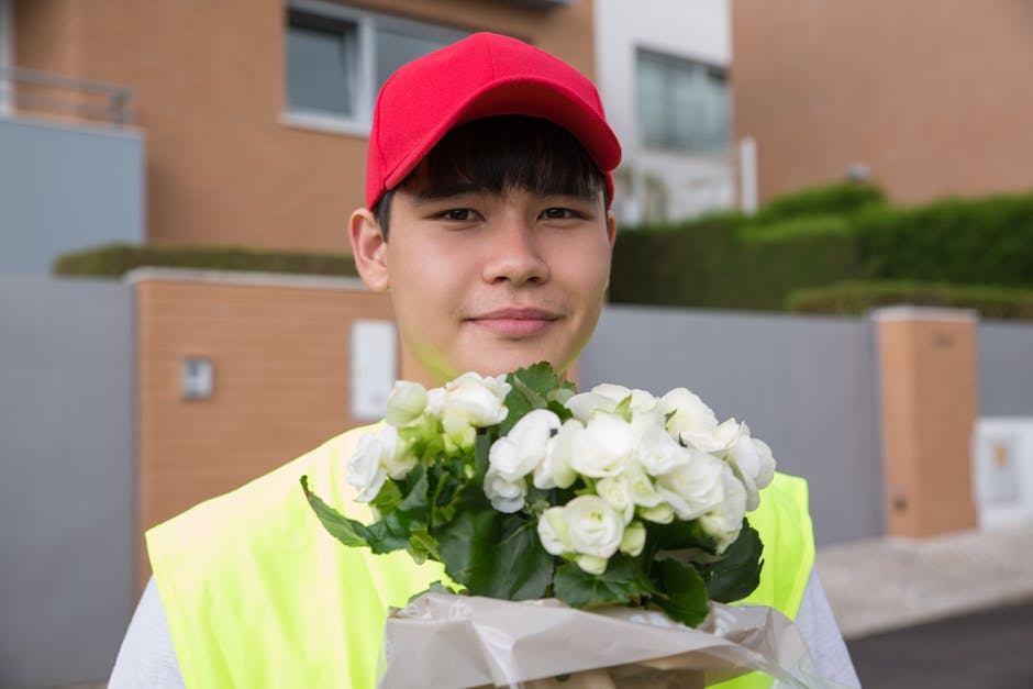 A delivery person wearing a bright yellow high-visibility vest and red cap holds a bouquet of fresh white roses wrapped in brown paper, smiling gently at the camera. The bouquet features tightly clust